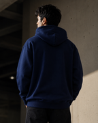 A man viewed from behind wearing a navy blue pullover hoodie and dark pants, standing in a concrete structure with dramatic side lighting.